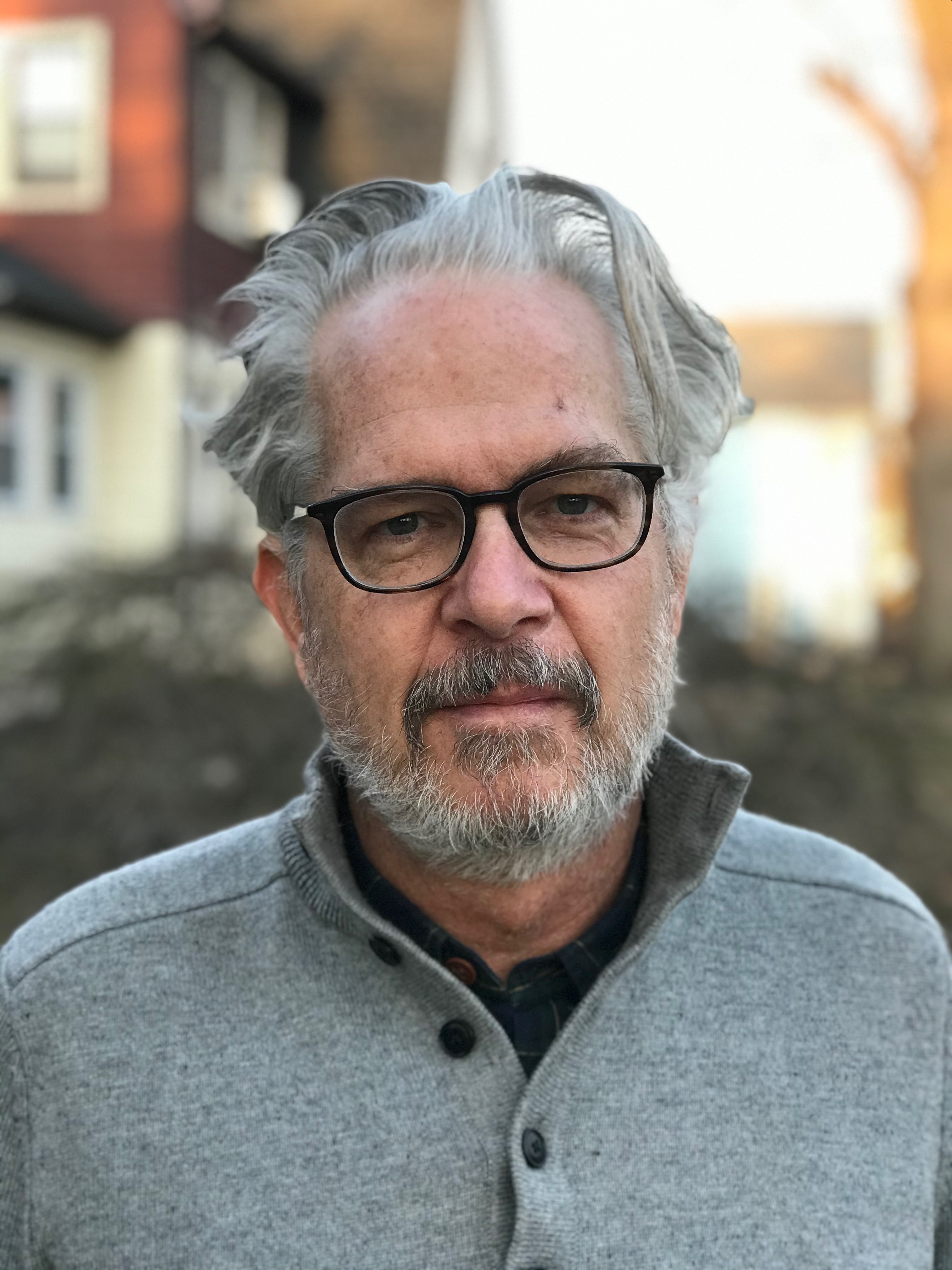 a man with grey hair and black framed glasses is standing outside looking at the camera.